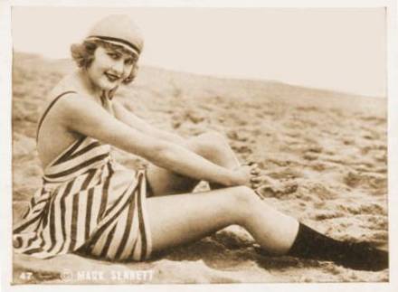 arcade-card-mack-sennett-comedies-woman-in-stripped-bathing-suit-and-cap-sitting-on-beach-one-knee-up-1920s